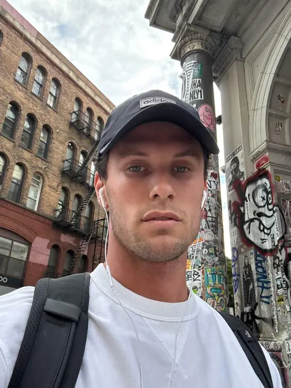 A young man in a cap and white T-shirt on a city street, looking to camera.